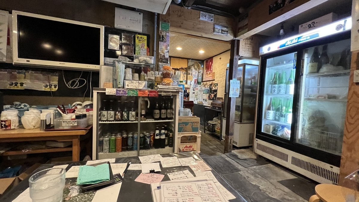 店内奥の冷蔵庫と酒類が並ぶ棚。地酒やビールが陳列された店内の一角。奥にはカウンター席も見える