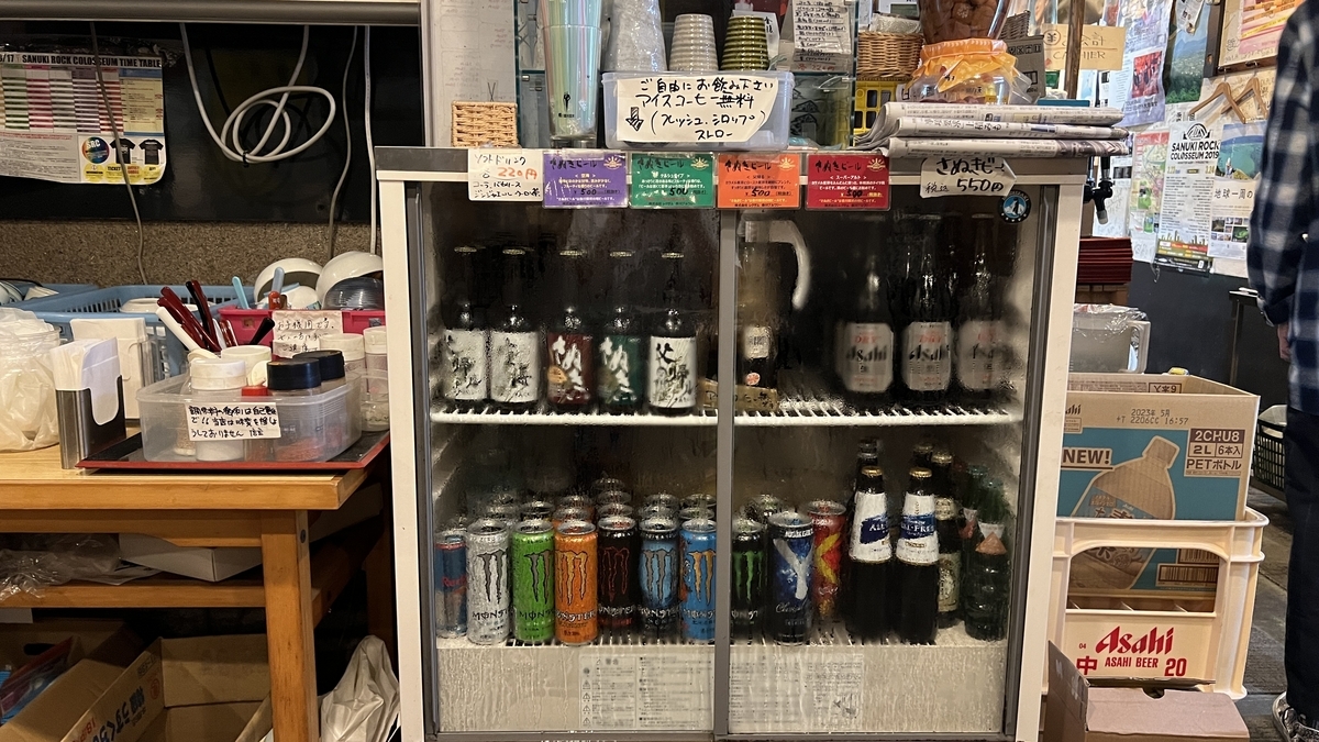 瓶ビールやソフトドリンクが並ぶ冷蔵ケース。上段には手書きの案内札や調味料が並ぶ