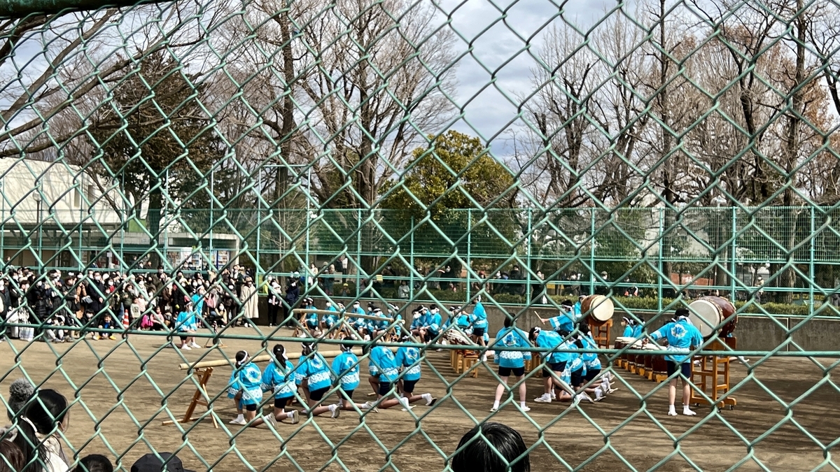 Children performing taiko drums behind a fence with audience watching