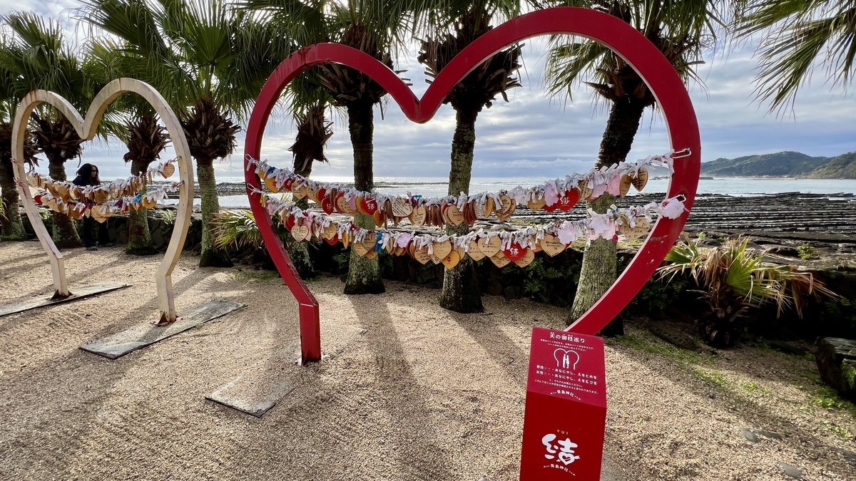 赤いハート型の枠に絵馬が掛けられた天の御柱めぐりの様子 背後に青島の海が広がる