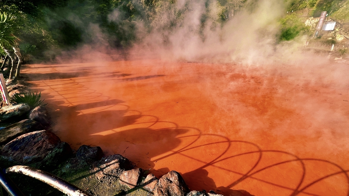 Vapor ascendiendo de la piscina de barro caliente marrón rojizo con sombras de barandillas en la superficie del agua en Chinoike Jigoku