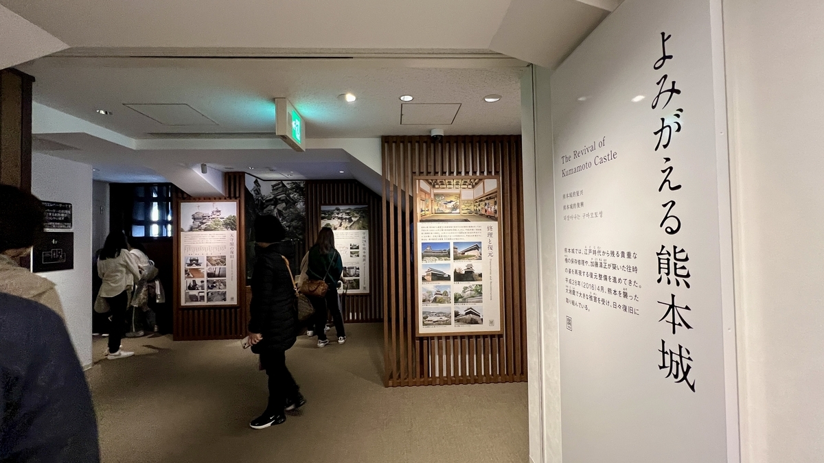 되살아나는 구마모토성 전시 입구에 서 있는 해설 패널과 복구 사진을 견학하는 방문객들