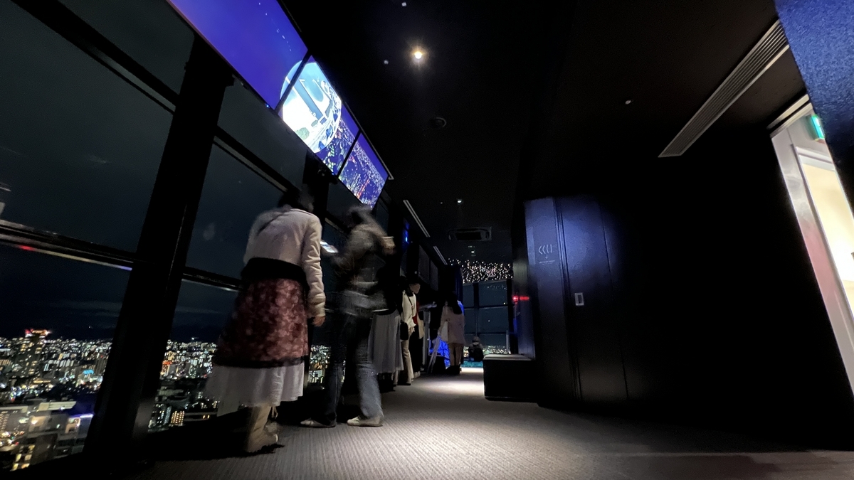 Visitors enjoying the night view at Fukuoka Tower observation deck with display panels by the windows