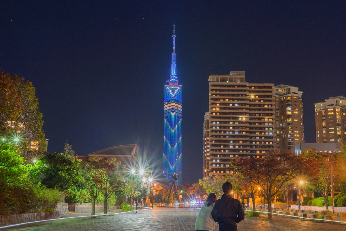 Fukuoka Tower glowing with blue illuminations and visitors strolling along the tree-lined path at night in Seaside Momochi