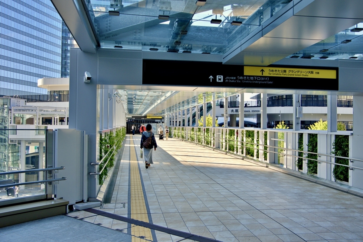 ผู้คนเดินผ่านทางเดินเชื่อมหลังคากระจก พร้อมป้ายบอกทางไปทางออกใต้ดิน Umekita ของสถานี JR Osaka