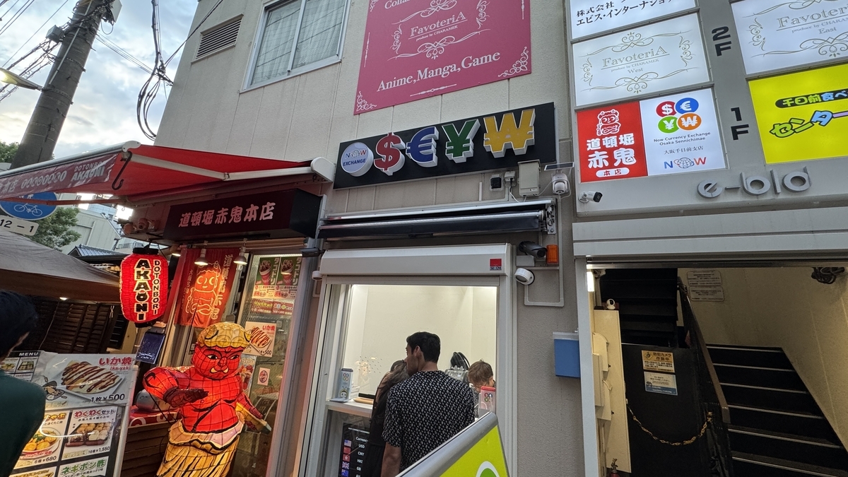 นักท่องเที่ยวกำลังแลกเงินที่ NOW CURRENCY EXCHANGE สาขาโอซาก้า Sennichimae ป้ายของร้าน Akako Honten อยู่ข้าง ๆ เคาน์เตอร์