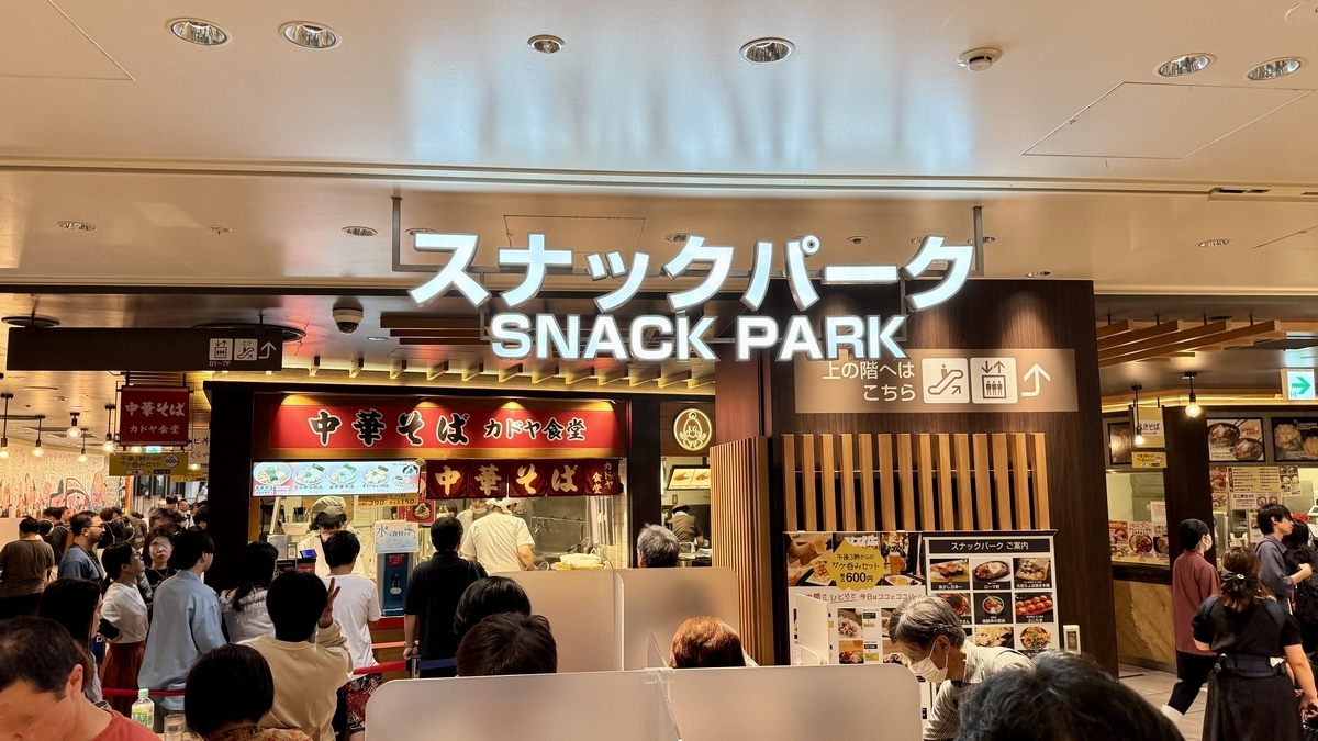 阪神梅田本店地下1階スナックパークの入口。白いロゴ看板の下に多くの人が集まり賑わう様子