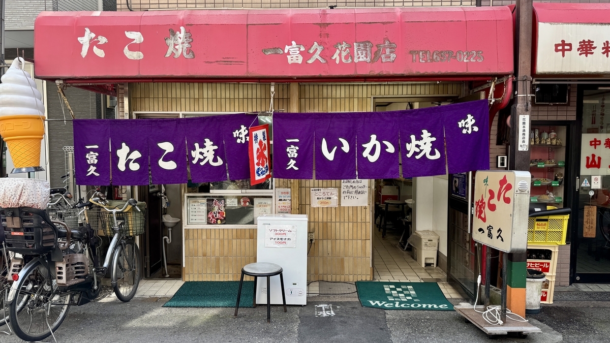 Exterior of Ichifuku with a purple noren curtain and signs advertising takoyaki and ika-yaki