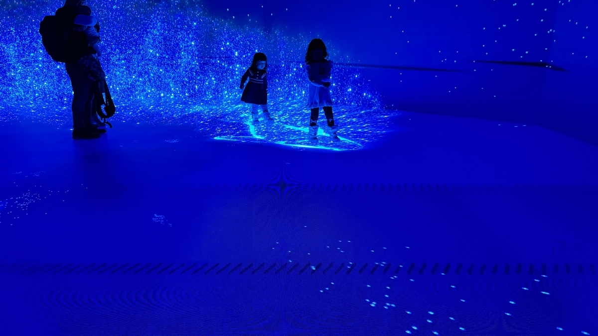 青い光の粒が広がる床で遊ぶ子どもと見守る大人の姿