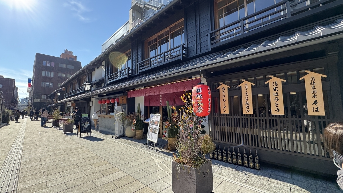 제등과 주홍색 노렌이 걸린 마치야 음식점, 고슈 호토 표지판이 늘어선 낮의 돌바닥 거리