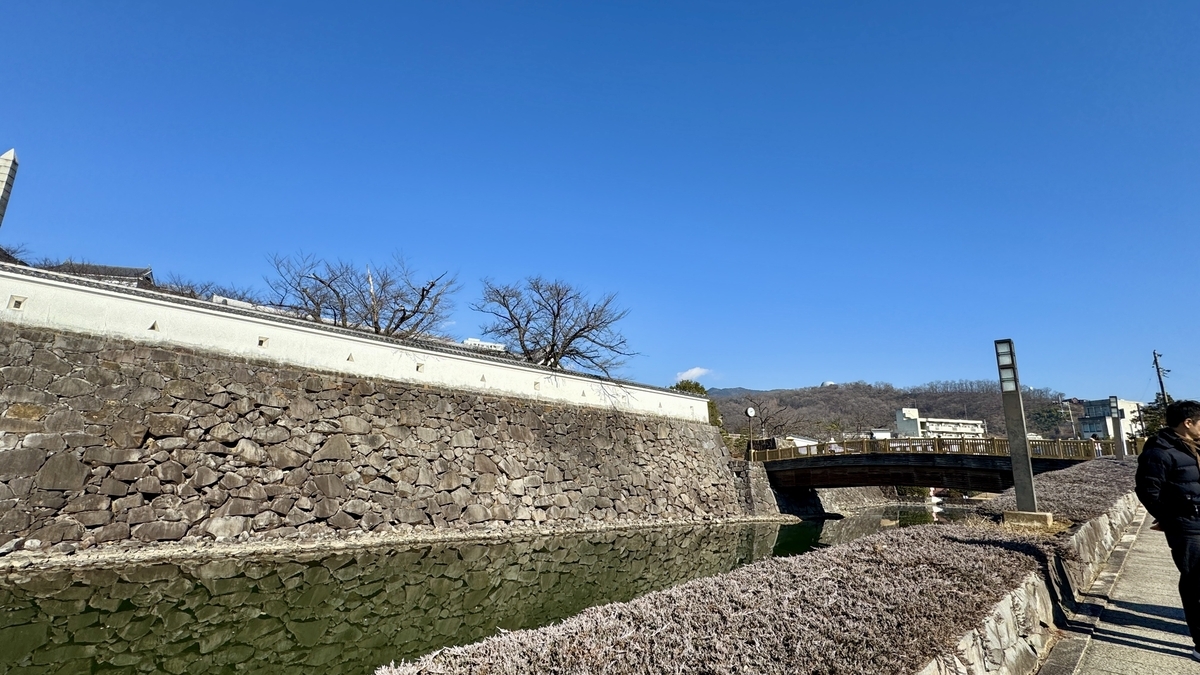 고후성 석축과 해자 수면이 이어지는 풍경, 푸른 하늘 아래 다리와 산책로가 뻗어 있는 낮 풍경