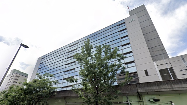 Exterior del edificio de la Sede del Area Metropolitana de JR Este con fachada de cristal reflejando bajo cielos azules y arboles de la calle