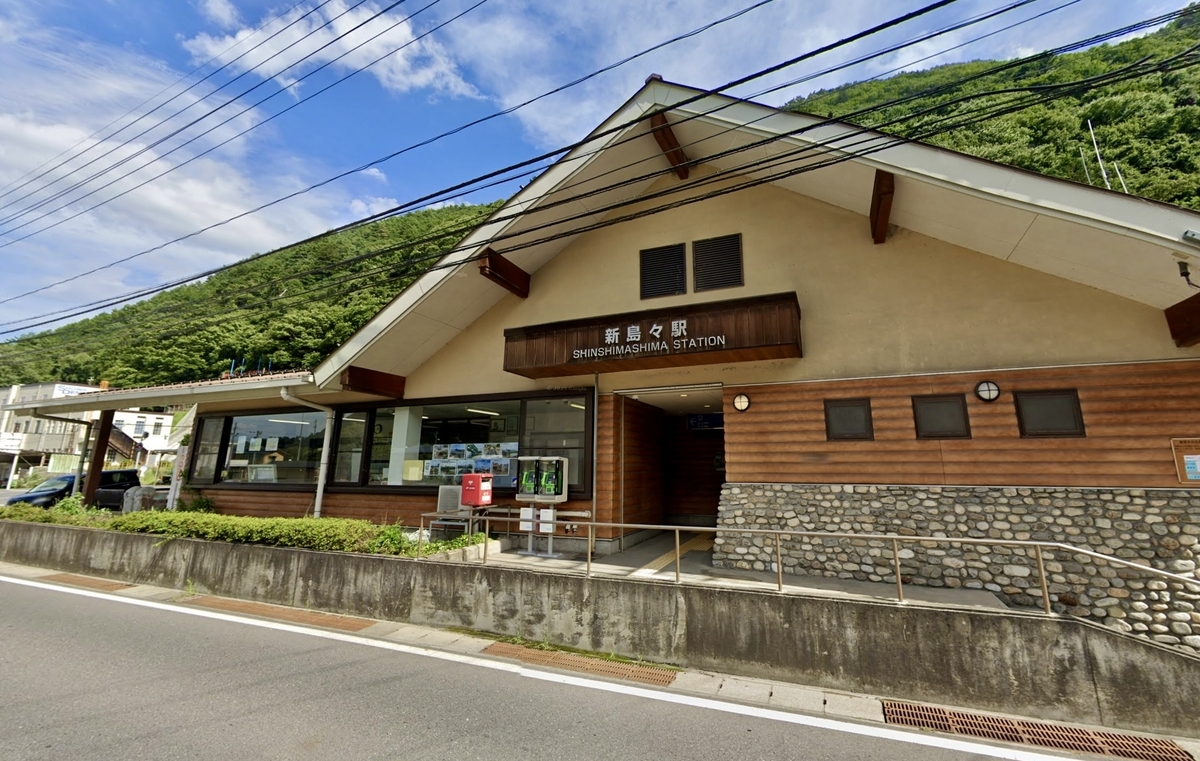 Edificio de estacion de estilo madera de la Estacion Shin-Shimashima con crestas de montanas verdes al fondo y letrero con el nombre de la estacion en la entrada