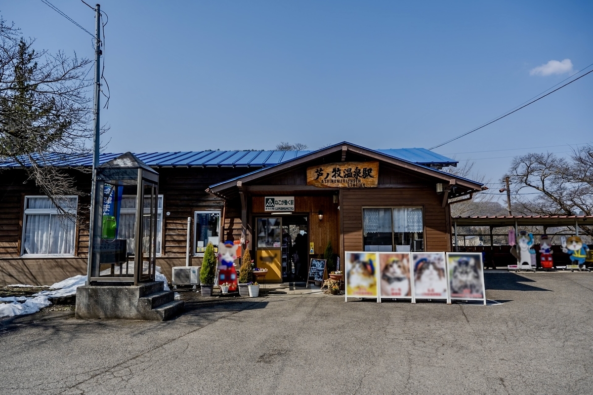 Edificio de estacion de madera con techo azul de la Estacion de Ashinomaki Onsen con un letrero de la estacion y paneles de fotos de gatos en la entrada