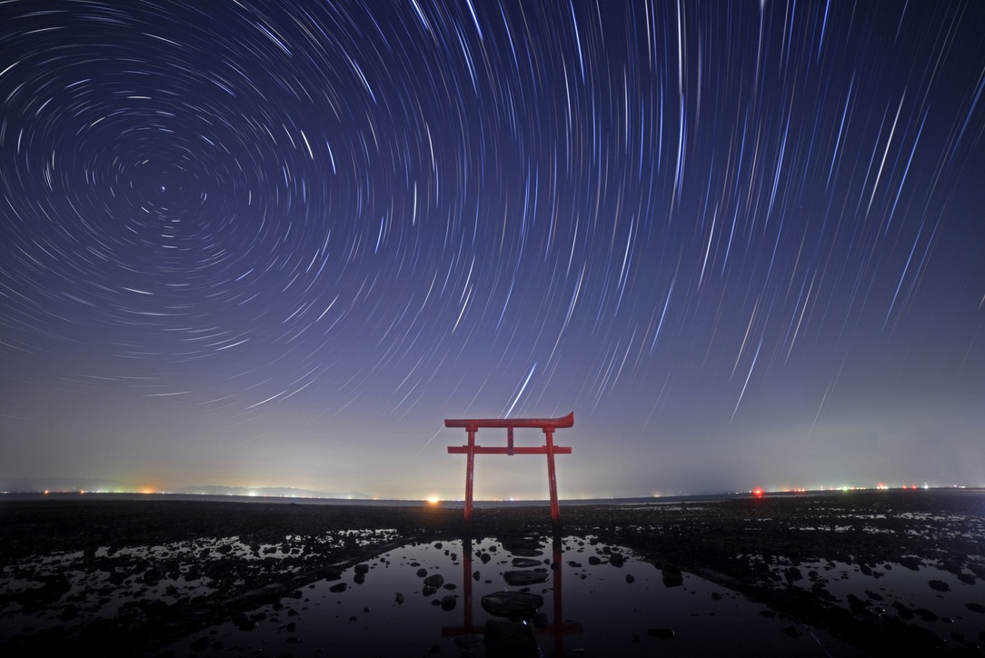 Torii rojo de pie en marismas reflejado en la superficie del agua bajo un cielo nocturno con rastros de estrellas