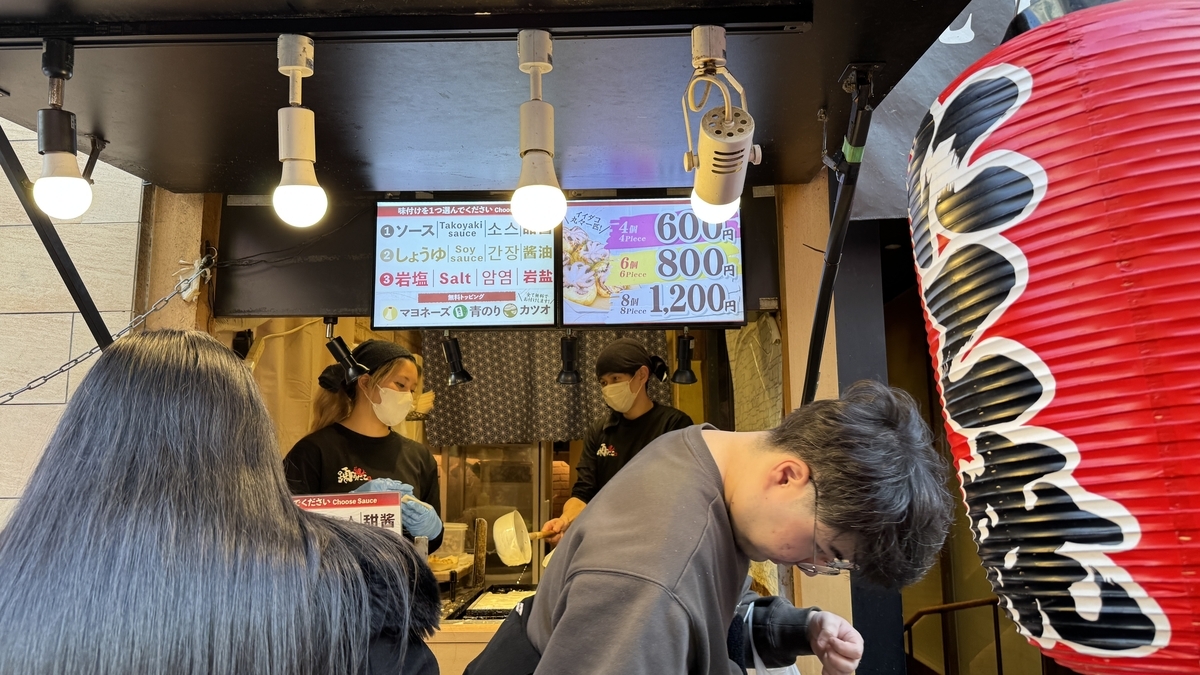 Staff grilling takoyaki at Odoridako storefront with flavor and price signs visible