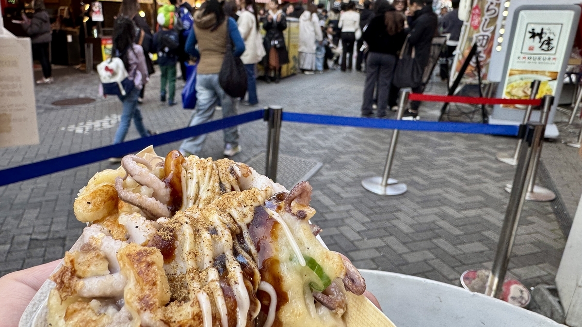 Hand holding Odoridako takoyaki with Dotonbori queue and rope barriers visible in background