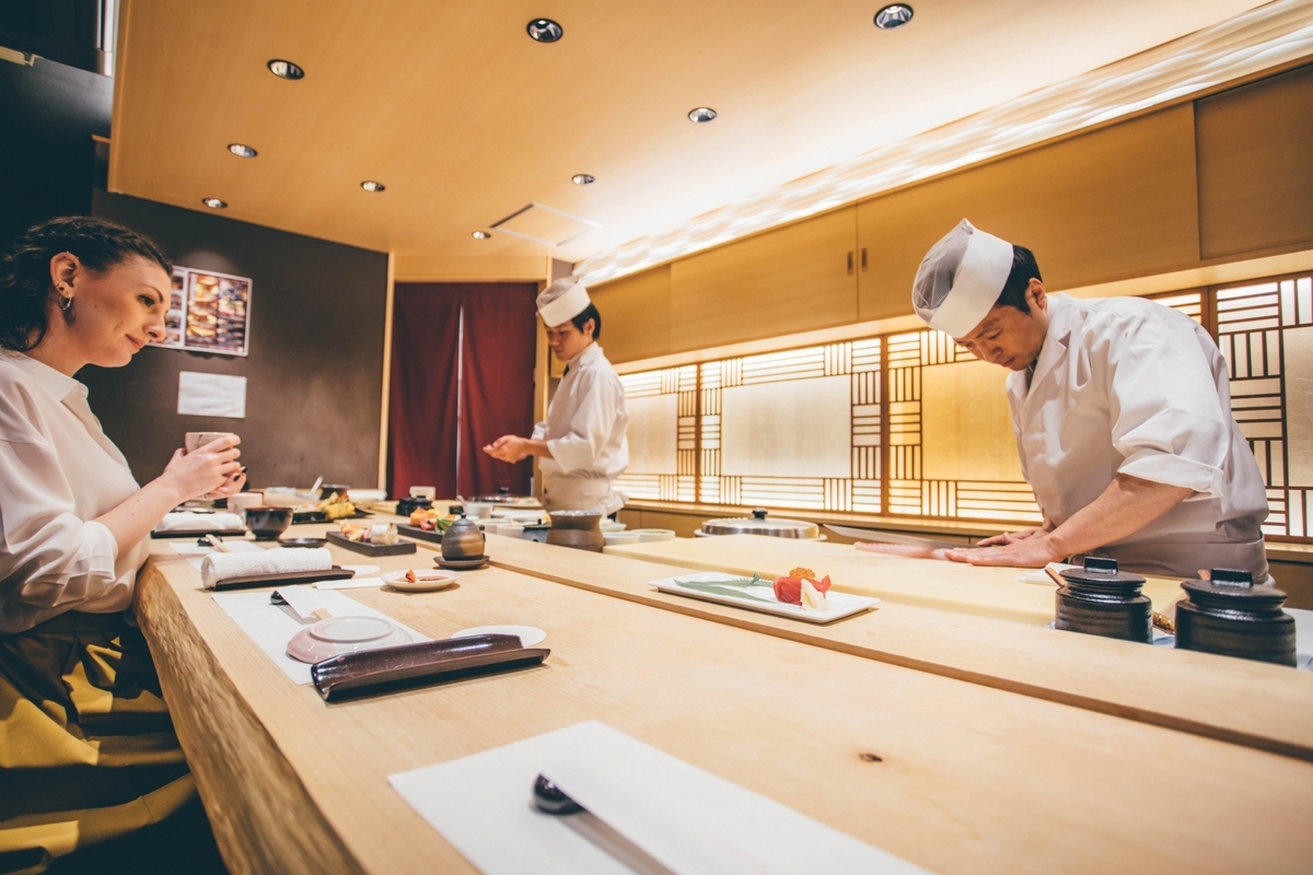 A sushi chef in traditional Japanese attire maintains a knife at a wooden counter while customers sit in the shop