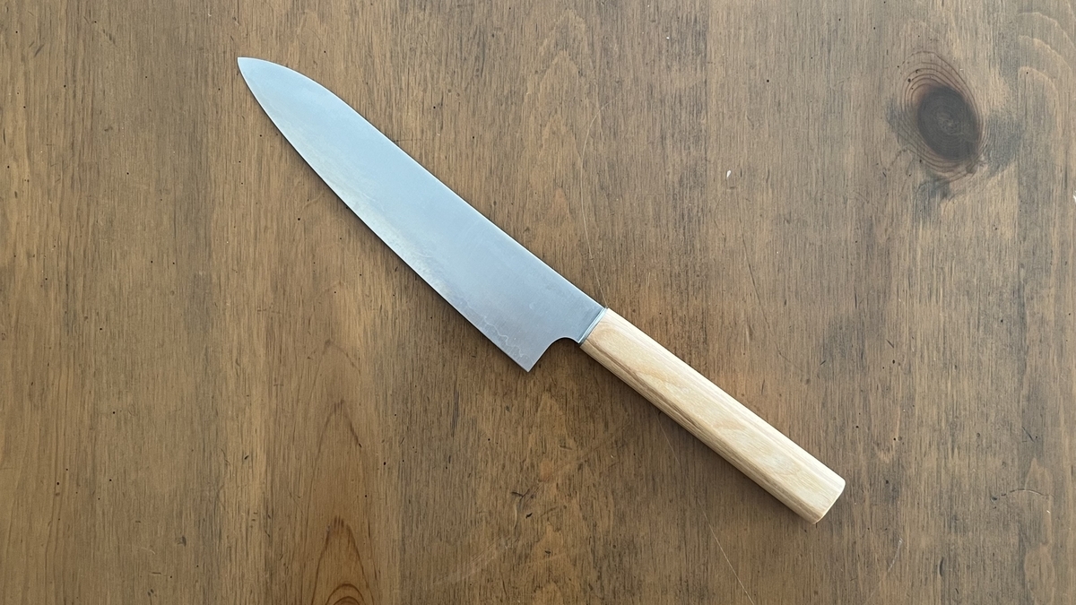 A Gyuto knife with a light wooden handle placed on a wood-grain table