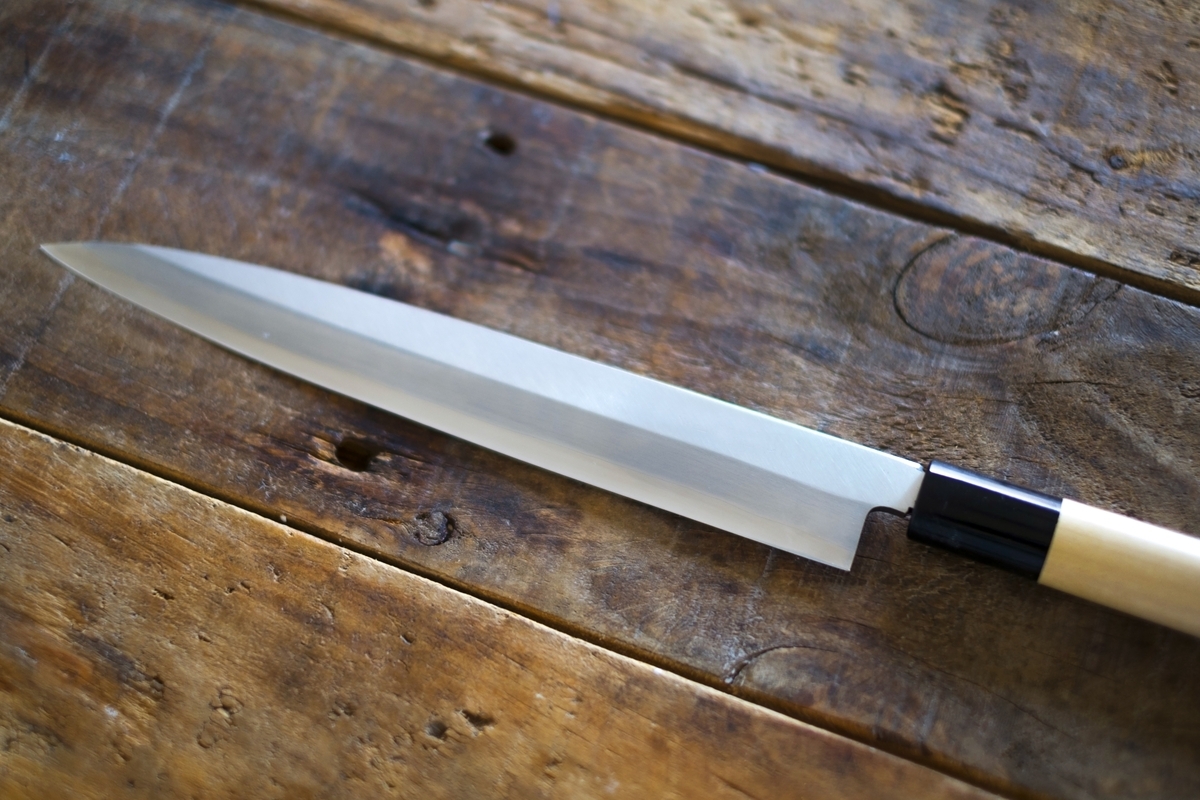 A long, slender Yanagiba knife with a single-bevel blade and white wooden handle on a wooden work surface