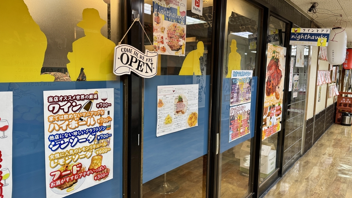面向新梅田食道街2樓走廊的nighthawks玻璃門和藍色下牆，黃色剪影和營業中招牌