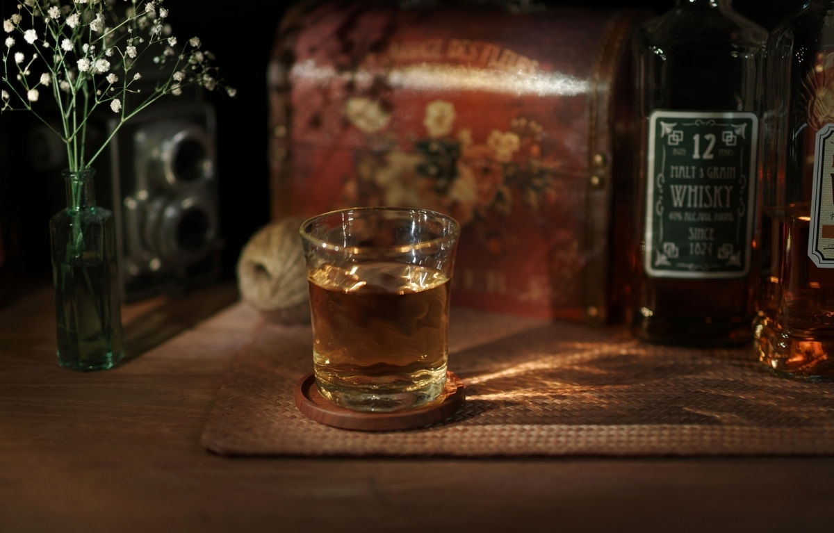 コースターの上のウイスキーグラスと周囲のボトルや小物を暖色光で写した卓上