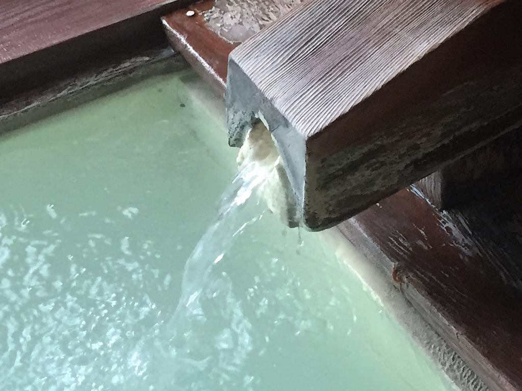Bluish, cloudy hot spring water flowing from a wooden spout into a tub